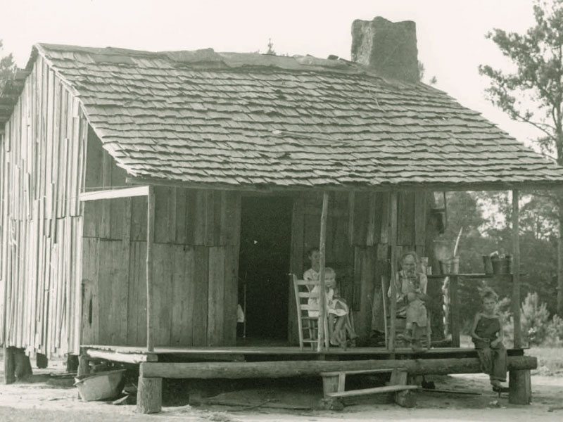 Black and white photograph of people sitting on a porch of a wooden house.